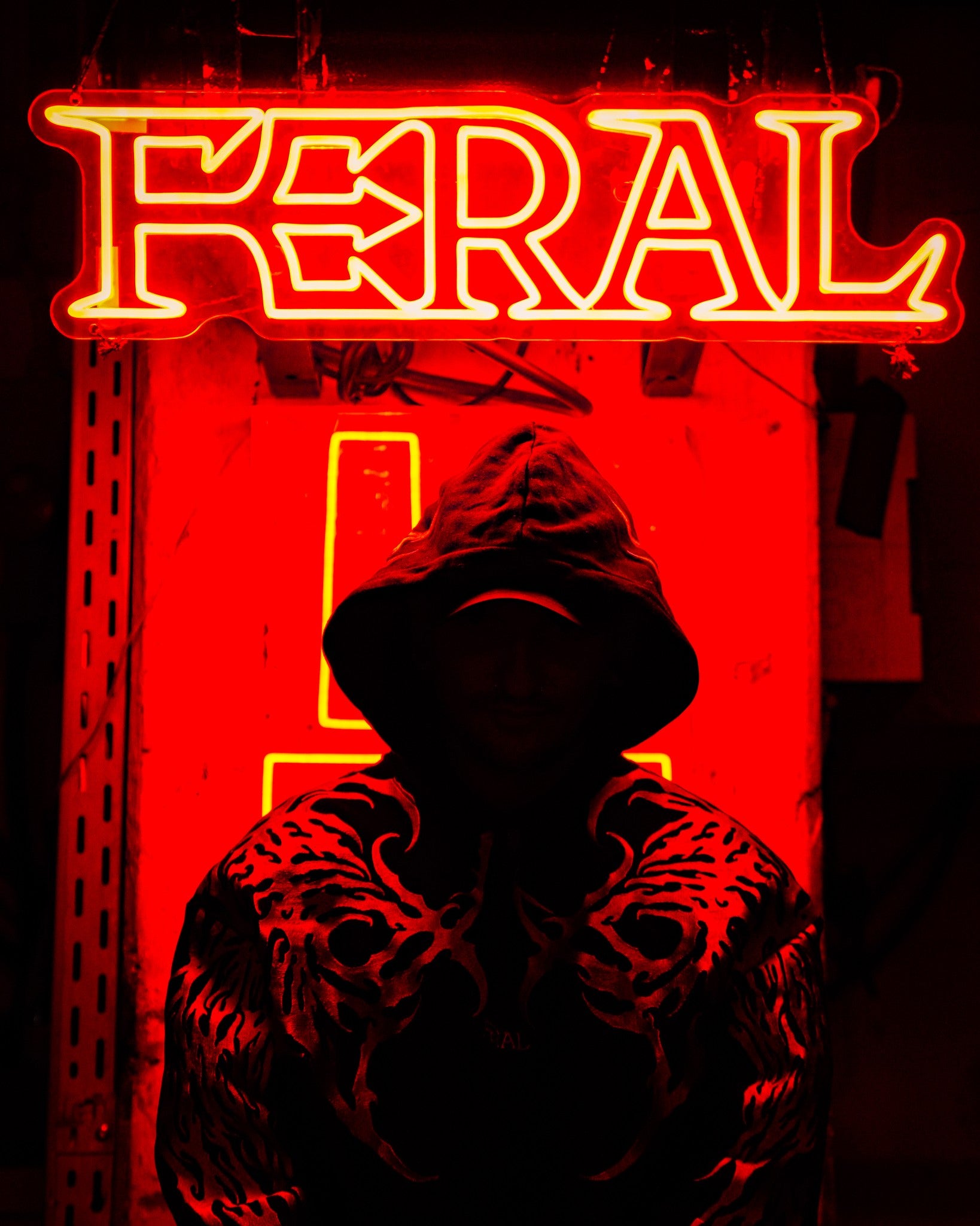 The FERAL neon sign hung above the DJ booth, raver in a black FERAL sigilism hoodie lit red beneath it